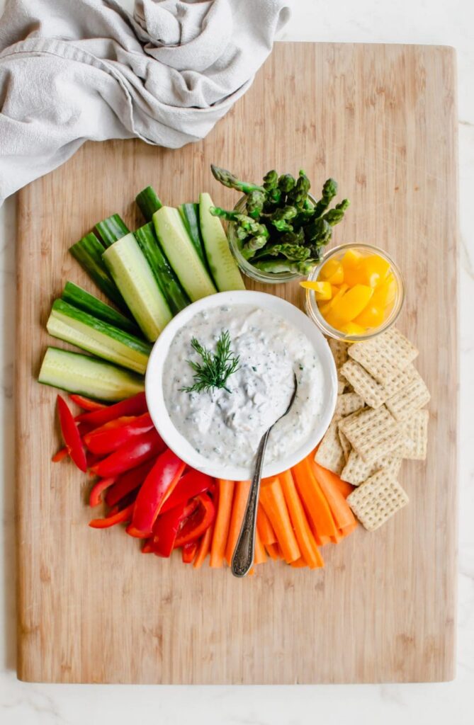Bowl of parmesan dip surrounded by sticks of veggies on a wooden board
